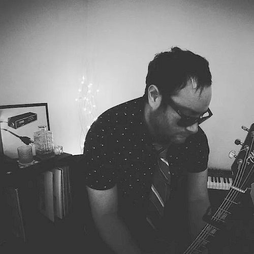 A man is playing a guitar in a dimly lit room with decorations, including a framed picture and various small items on a shelf.