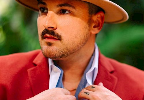 A person wearing a red blazer and hat is adjusting a scarf or fabric around their neck. They have a mustache and the background is out of focus.