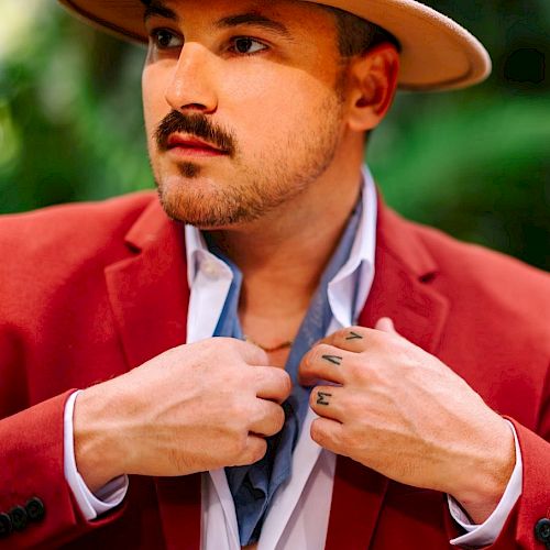 A person wearing a red blazer and hat is adjusting a scarf or fabric around their neck. They have a mustache and the background is out of focus.