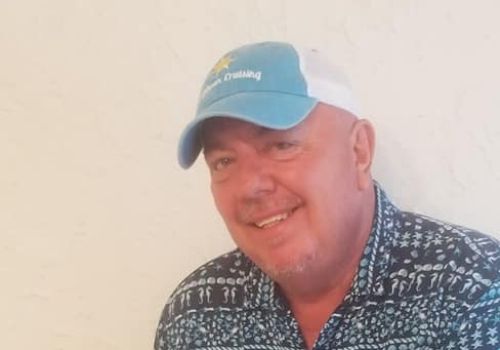 A person wearing a blue shirt and cap is smiling while holding a guitar in front of a light-colored textured wall.