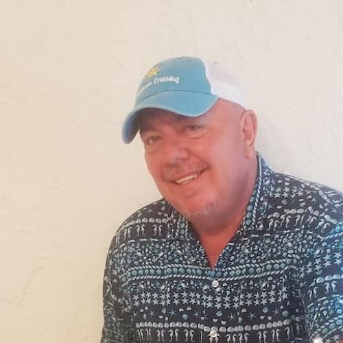 A person wearing a blue shirt and cap is smiling while holding a guitar in front of a light-colored textured wall.