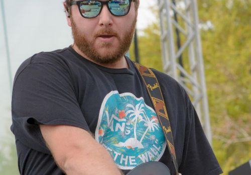 A person with sunglasses and a cap plays an electric guitar on stage at a Reggae Rise Up event, as indicated by the logo in the bottom corner.