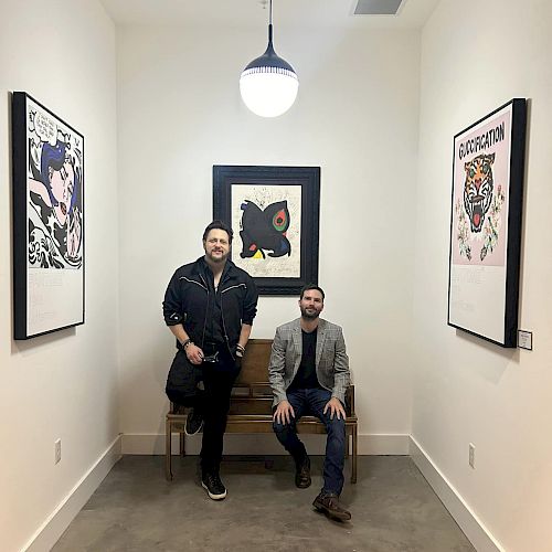 Two men in a narrow room, one standing and one sitting on a bench, with artwork on the walls and a hanging light above them.