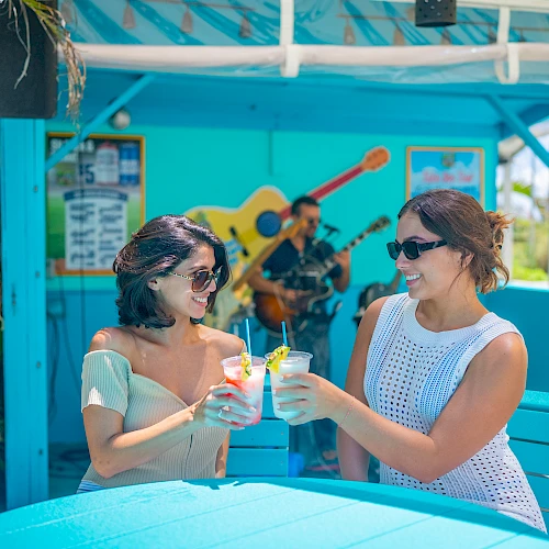 Two people are clinking glasses at an outdoor caf&eacute; with a musician playing guitar in the background, surrounded by vibrant blue d&eacute;cor.