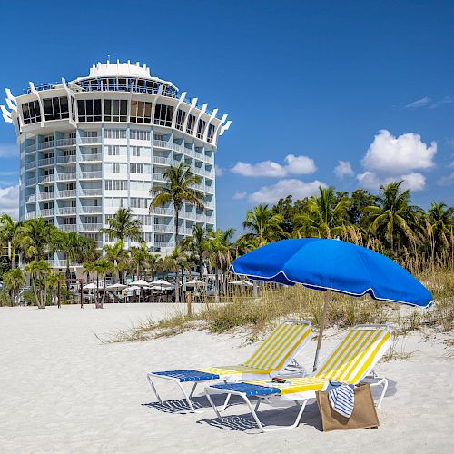 Two beach chairs with a blue umbrella sit on sandy beach; a tall, modern building and palm trees are visible in the background.