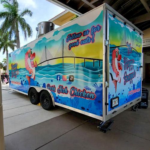 A colorful food truck with seaside graphics and text promoting fish and chicken dishes is parked by a building with palm trees.