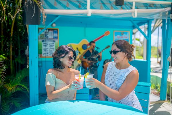 Two people are toasting drinks at an outdoor bar with a musician playing guitar in the background.