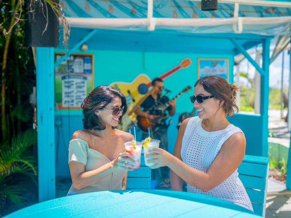Two people are toasting drinks at an outdoor bar with a musician playing guitar in the background.