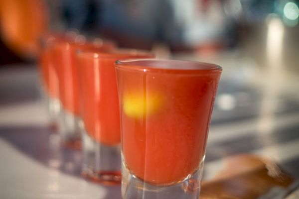 The image shows several small glasses filled with a red-orange liquid, likely a drink, aligned on a table in sunlight.