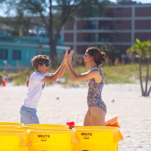 Two kids are playing on the beach, giving each other a high-five. Yellow bins are in the foreground, with buildings and trees behind them.