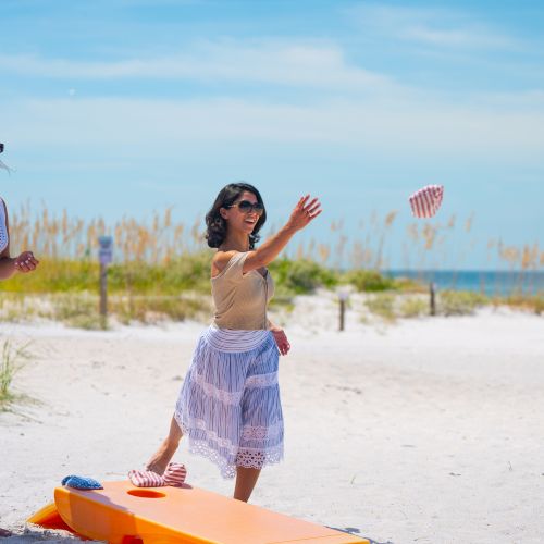 Two people enjoying a beach day, playing a tossing game with a board and corn bags, under a clear blue sky.