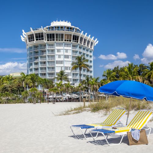 A beach scene with two loungers under a blue umbrella, palm trees, and a white multi-story building in the background.