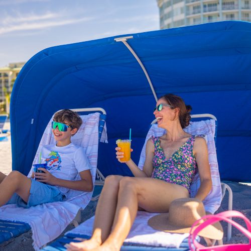 A woman and a child relax on beach loungers under a blue canopy, enjoying drinks.