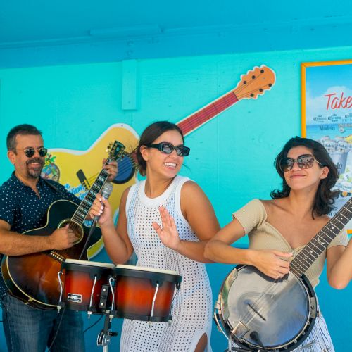 A group of three people are playing instruments in a colorful setting with guitars and promotional signs in the background.