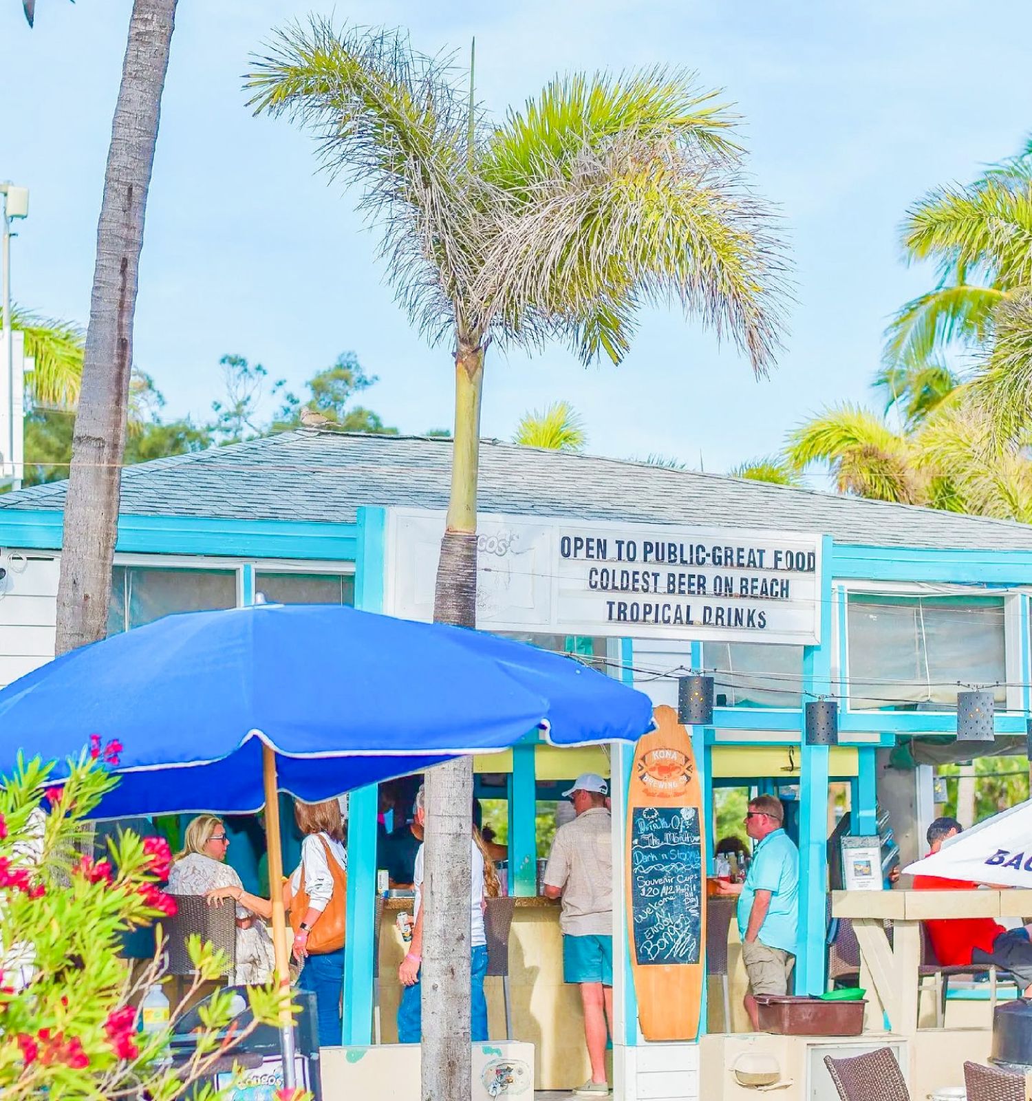 This image features an outdoor beachside cafe with people gathered around. There are palm trees, a blue umbrella, and a sign on the building.